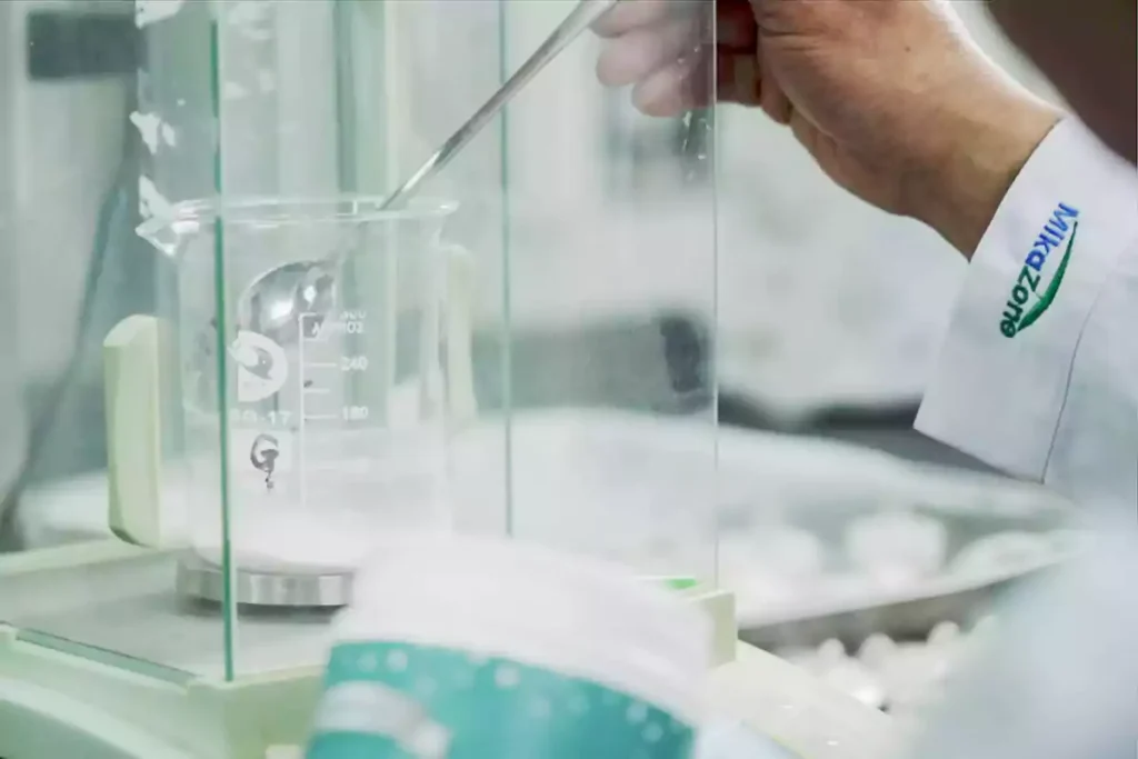 A person wearing a white lab coat with the Mikazone logo is pouring white powder into a beaker.