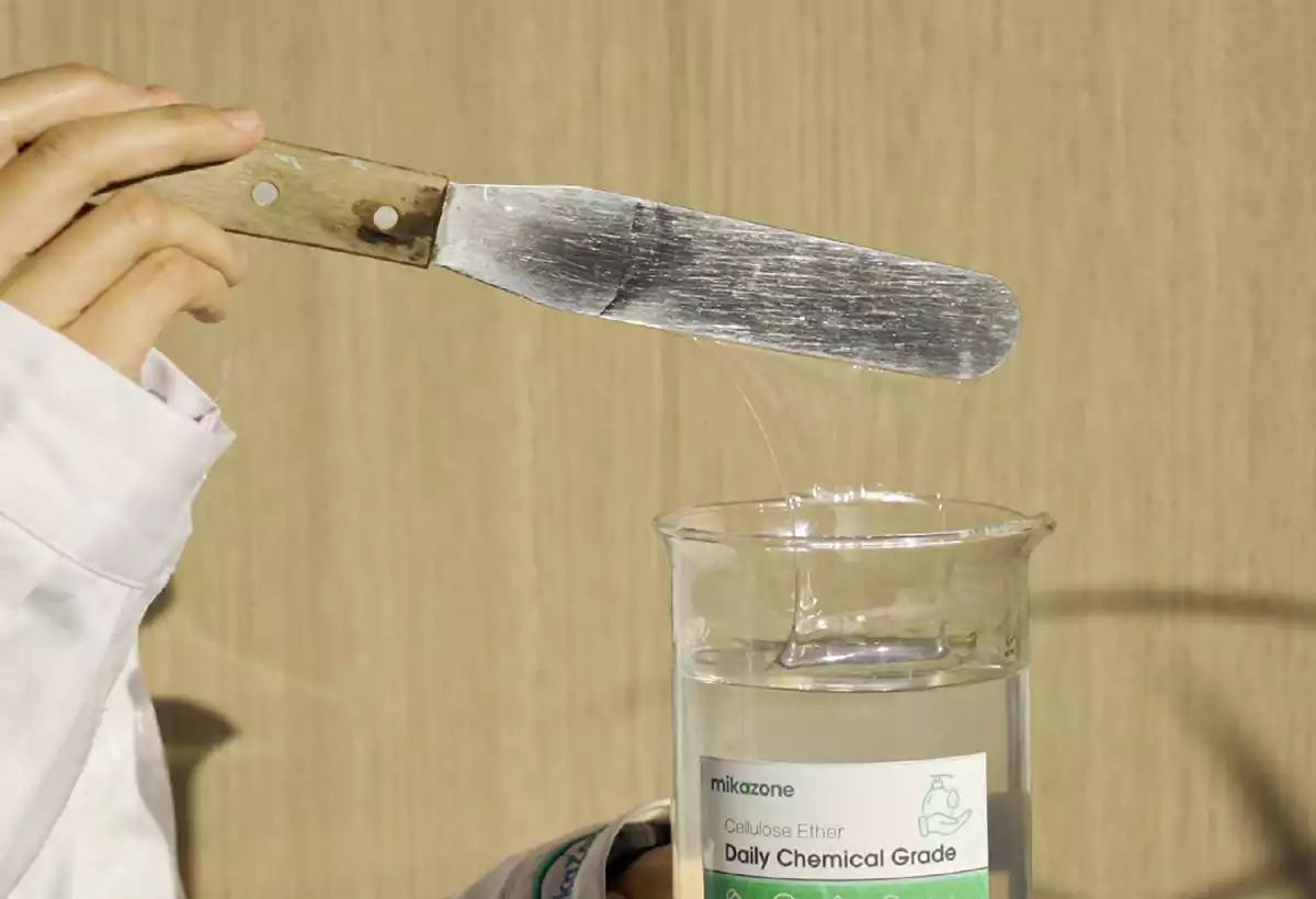 A person in a lab coat uses a spatula to lift a cosmetic-grade cellulose ether solution to demonstrate its transparency and viscosity.