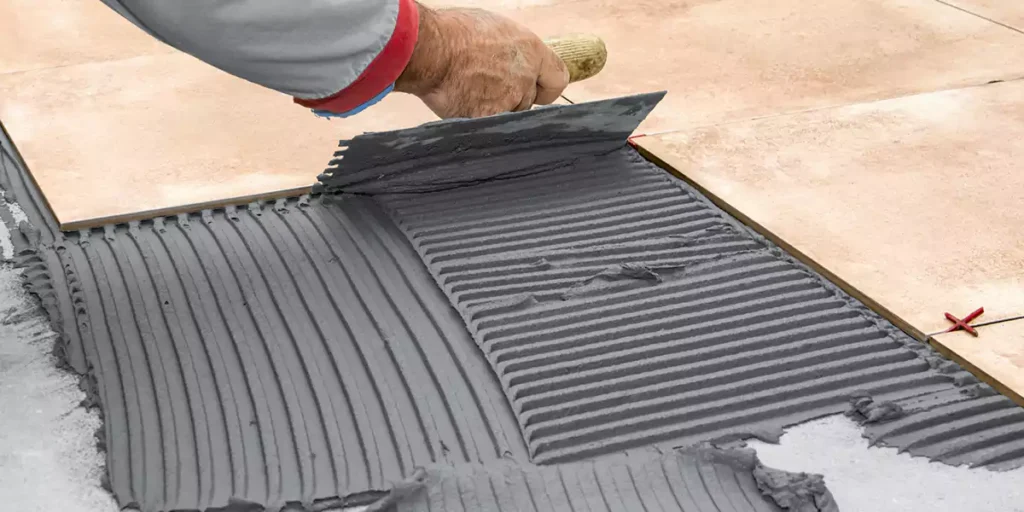 A worker using a serrated trowel to apply gray tile adhesive on the cement floor.